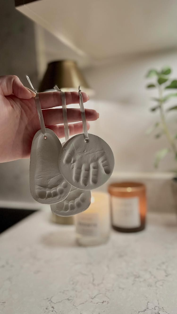 Hand holding two grey salt dough ornaments featuring a child's footprint and handprint with white ribbons.
