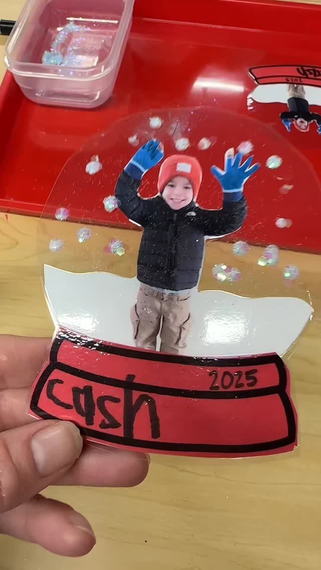 A hand holds a handmade photo ornament with sequins and a red base labeled Cash 2025.