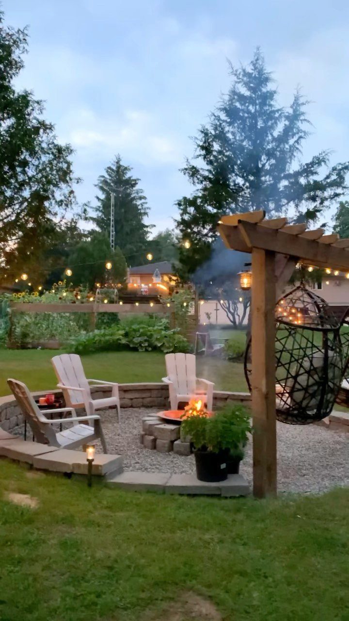 A stone fire pit with a burning fire surrounded by white Adirondack chairs in a backyard.