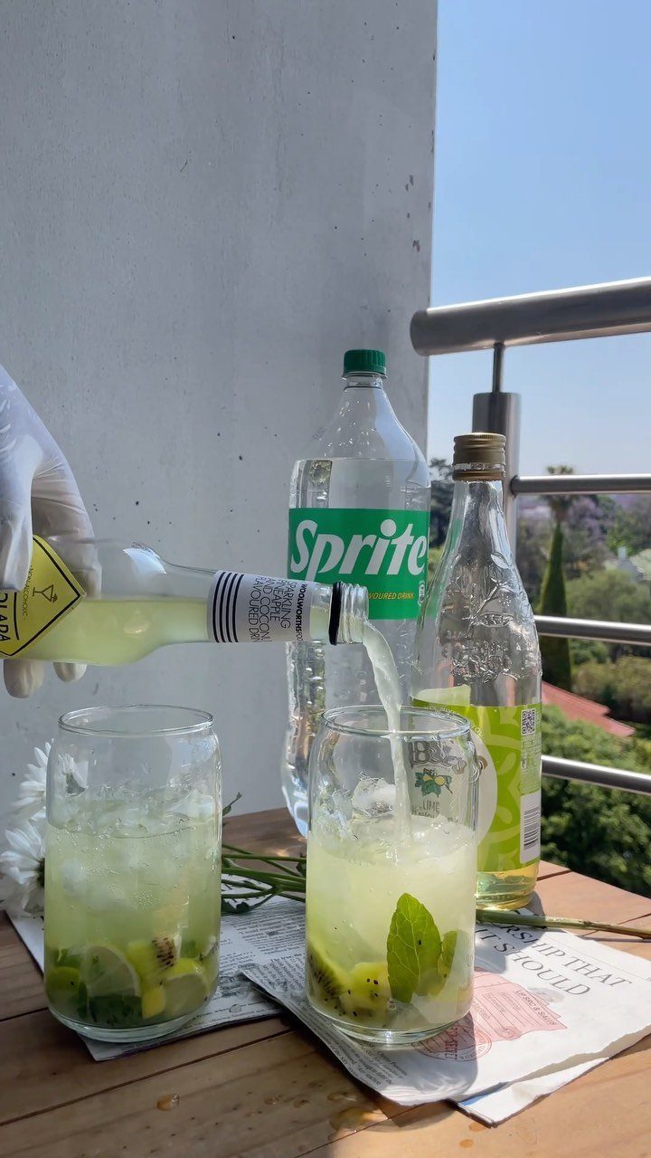 A gloved hand pouring liquid from a bottle into a glass of fruit-infused iced drink.
