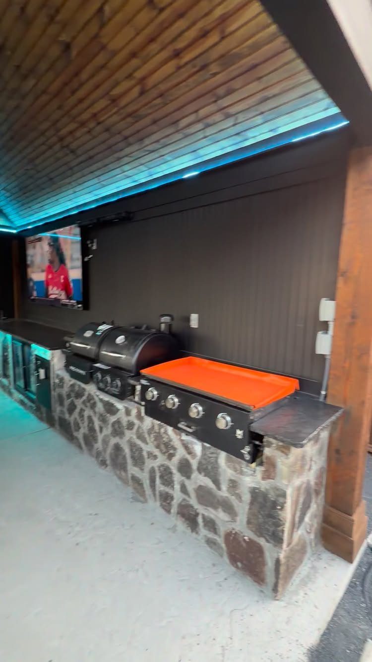 Built-in outdoor kitchen with stone base, black grill, orange griddle, and mounted television.