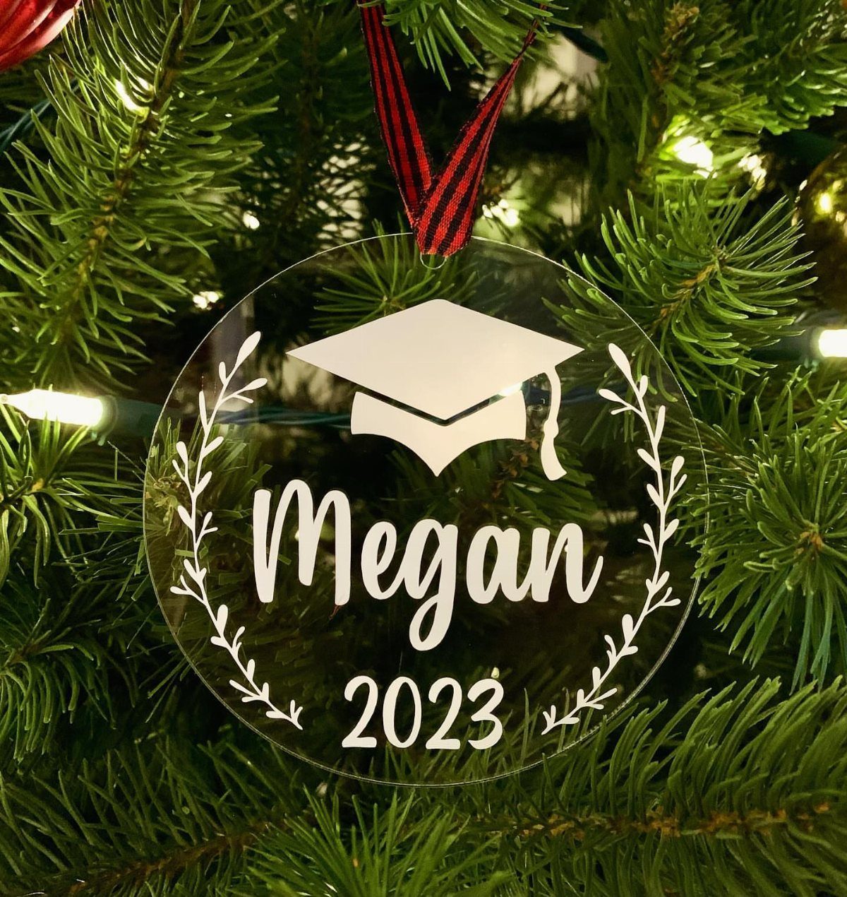 Clear acrylic graduation ornament with white text and cap icon hanging on a Christmas tree.