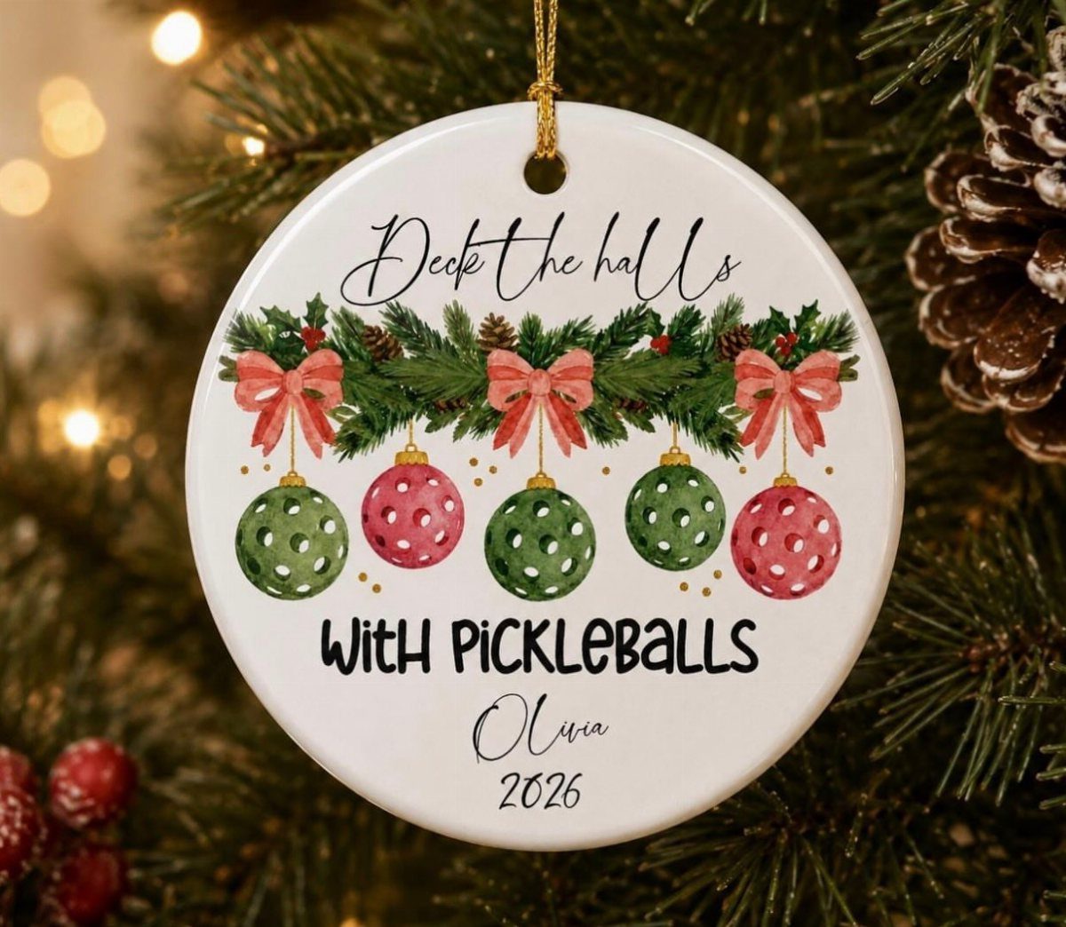 A white ceramic Christmas ornament with pickleball themed text hanging on a pine tree branch.