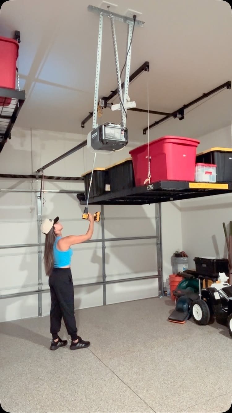 6 Small Garage Building Ideas for Storage, Projects & Living Space 1 A woman installing a black overhead storage rack holding large plastic bins in a garage.