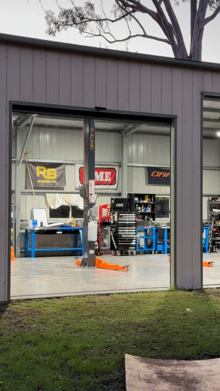 An open automotive workshop featuring a car lift, blue workbenches, and tool chests inside a grey metal building.