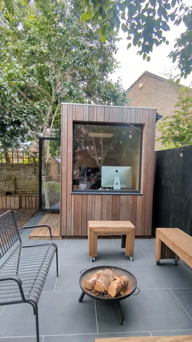 A modern wooden garden studio with a large window overlooking a tiled patio area.