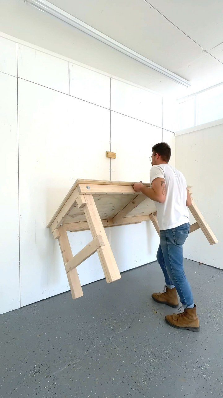 A man lifting a wooden workbench structure against a white wall in a workshop.