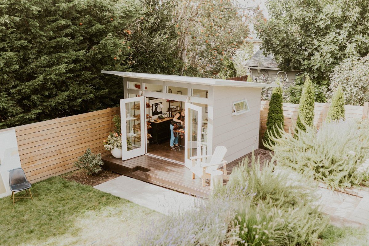 A modern white garden shed with open glass doors revealing a functional workshop interior.